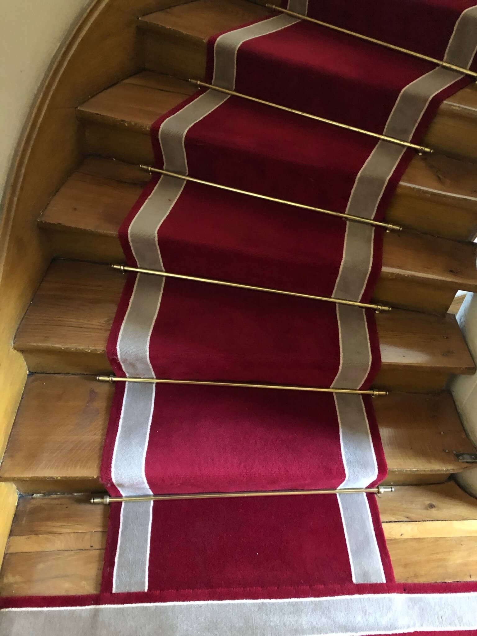 Escalier en bois avec un tapis rouge central bordé de blanc fixé par des barres dorées.