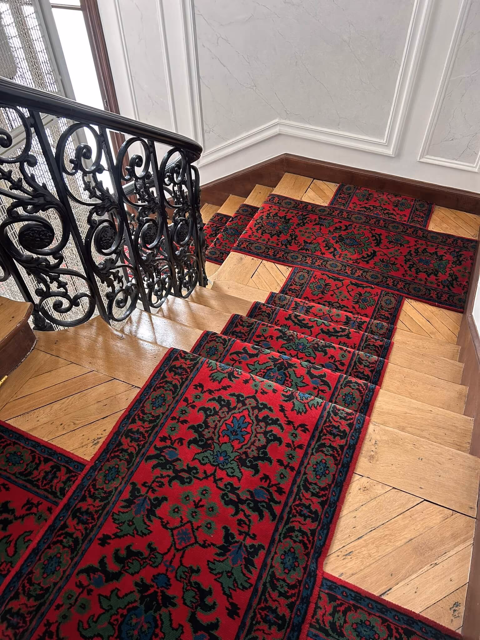 Escalier en bois avec un tapis rouge à motifs verts et noirs et une rampe en fer forgé noir.