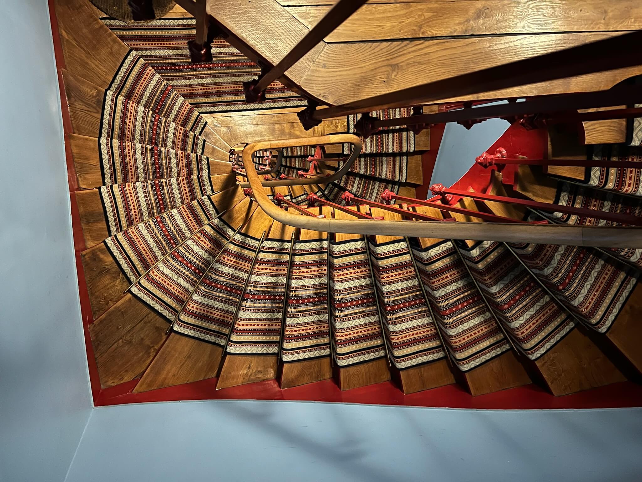 Vue en plongée d'un escalier en colimaçon en bois avec tapis à motifs rouges et blancs.