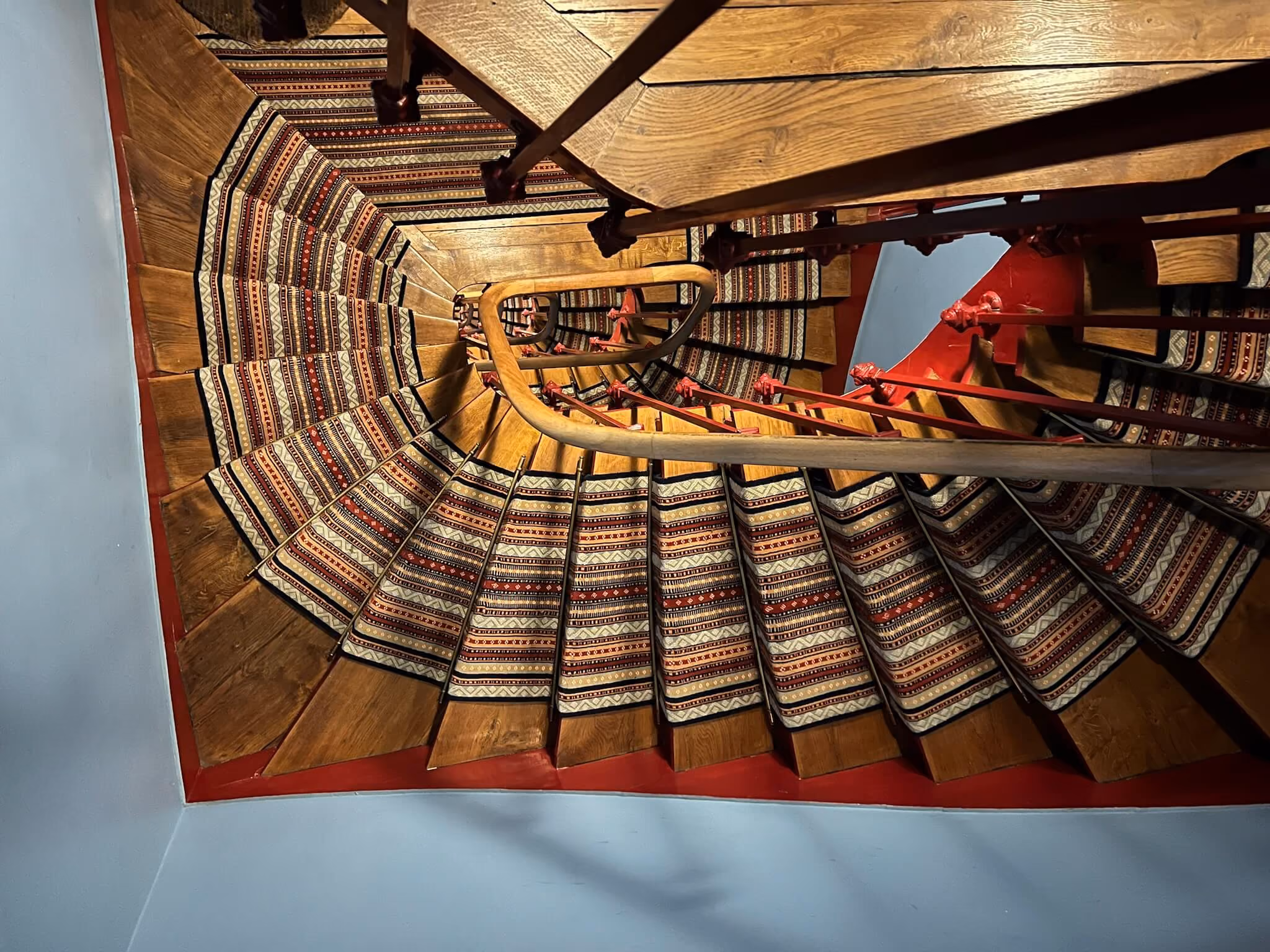 Vue en plongée d'un escalier en colimaçon en bois avec tapis à motifs rouges et blancs.