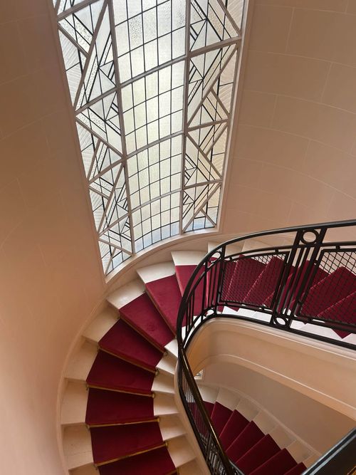 Escalier en colimaçon avec tapis rouge et rampe en fer noir, vitrail décoratif à côté.