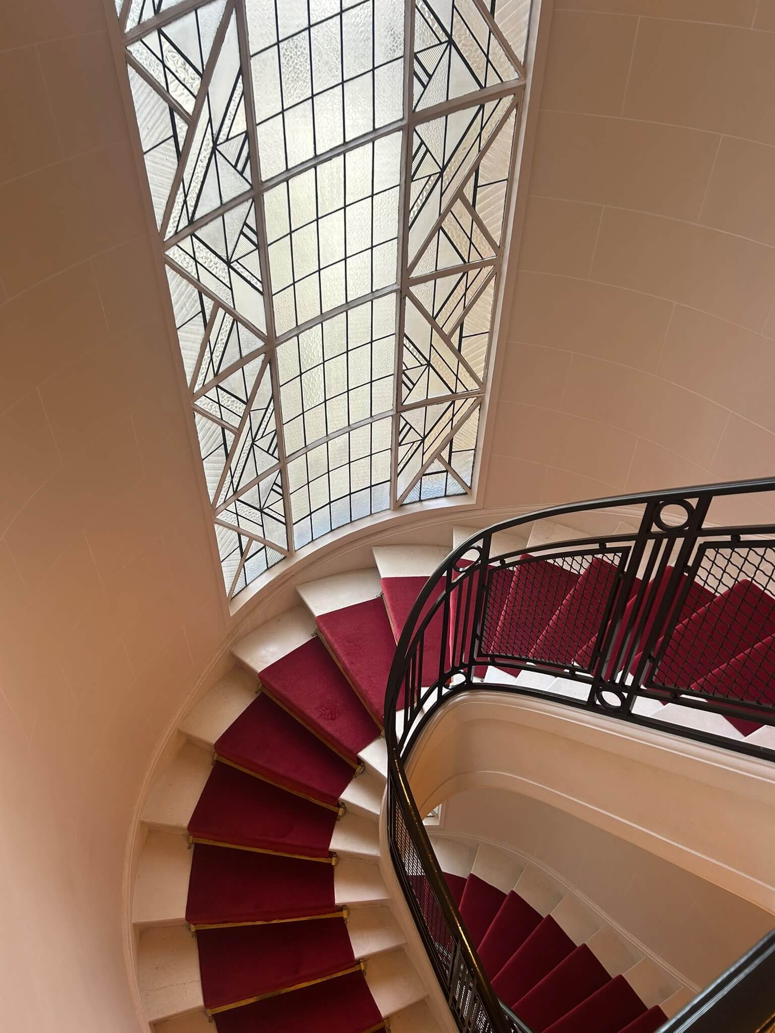 Escalier en colimaçon avec tapis rouge et rampe en fer noir, vitrail décoratif à côté.