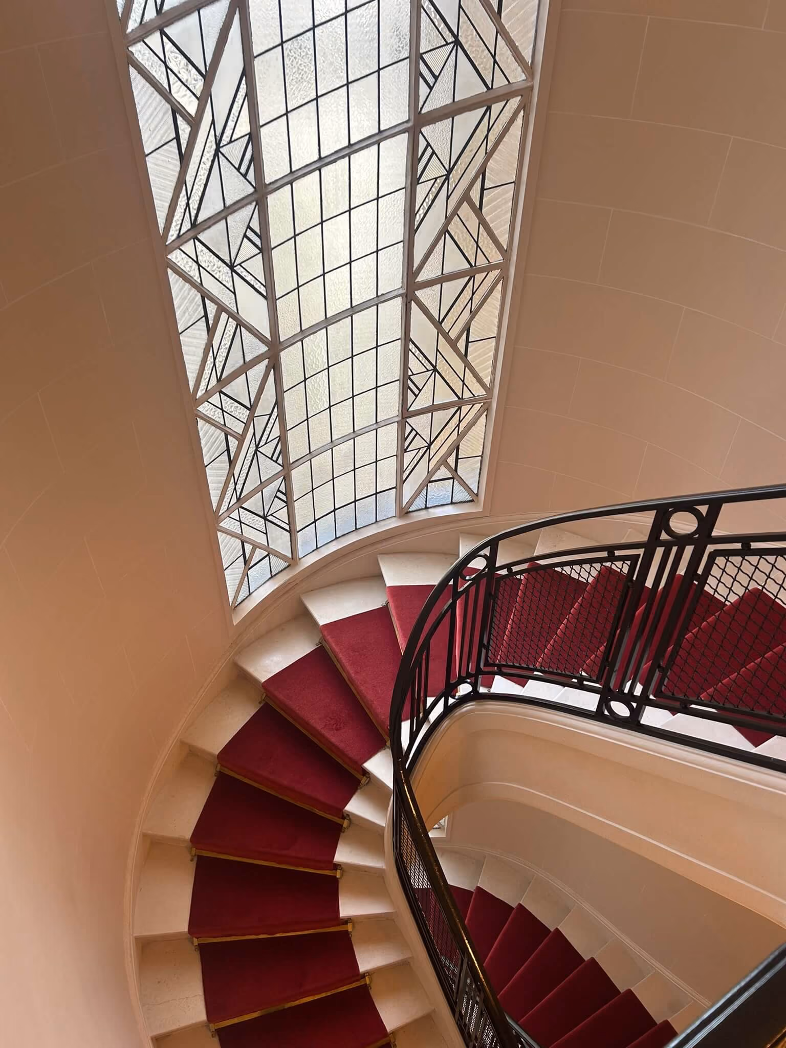 Escalier en colimaçon avec tapis rouge et rampe en fer noir, vitrail décoratif à côté.