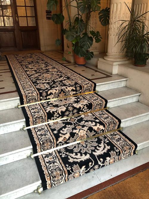 Escalier en marbre avec un tapis décoré noir et beige maintenu par trois barres dorées.