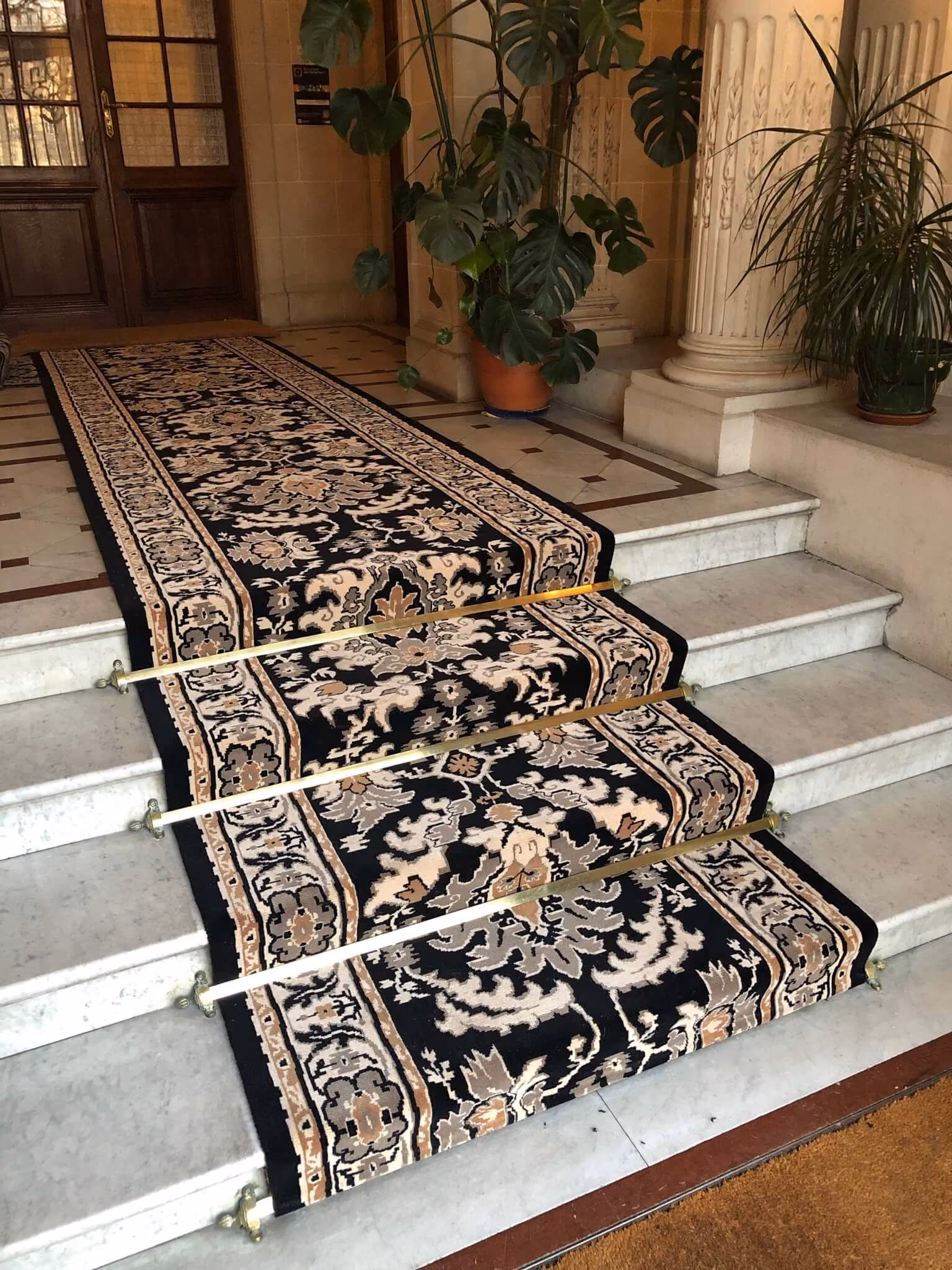Escalier en marbre avec un tapis décoré noir et beige maintenu par trois barres dorées.
