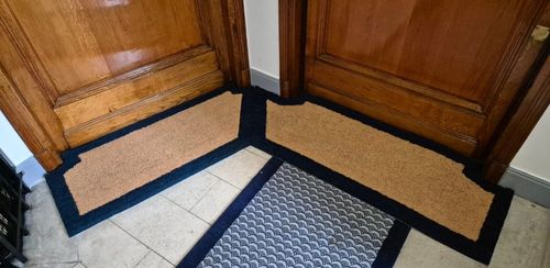 Deux paillassons d'entrée en coin avec cadre noir devant des portes en bois foncé.