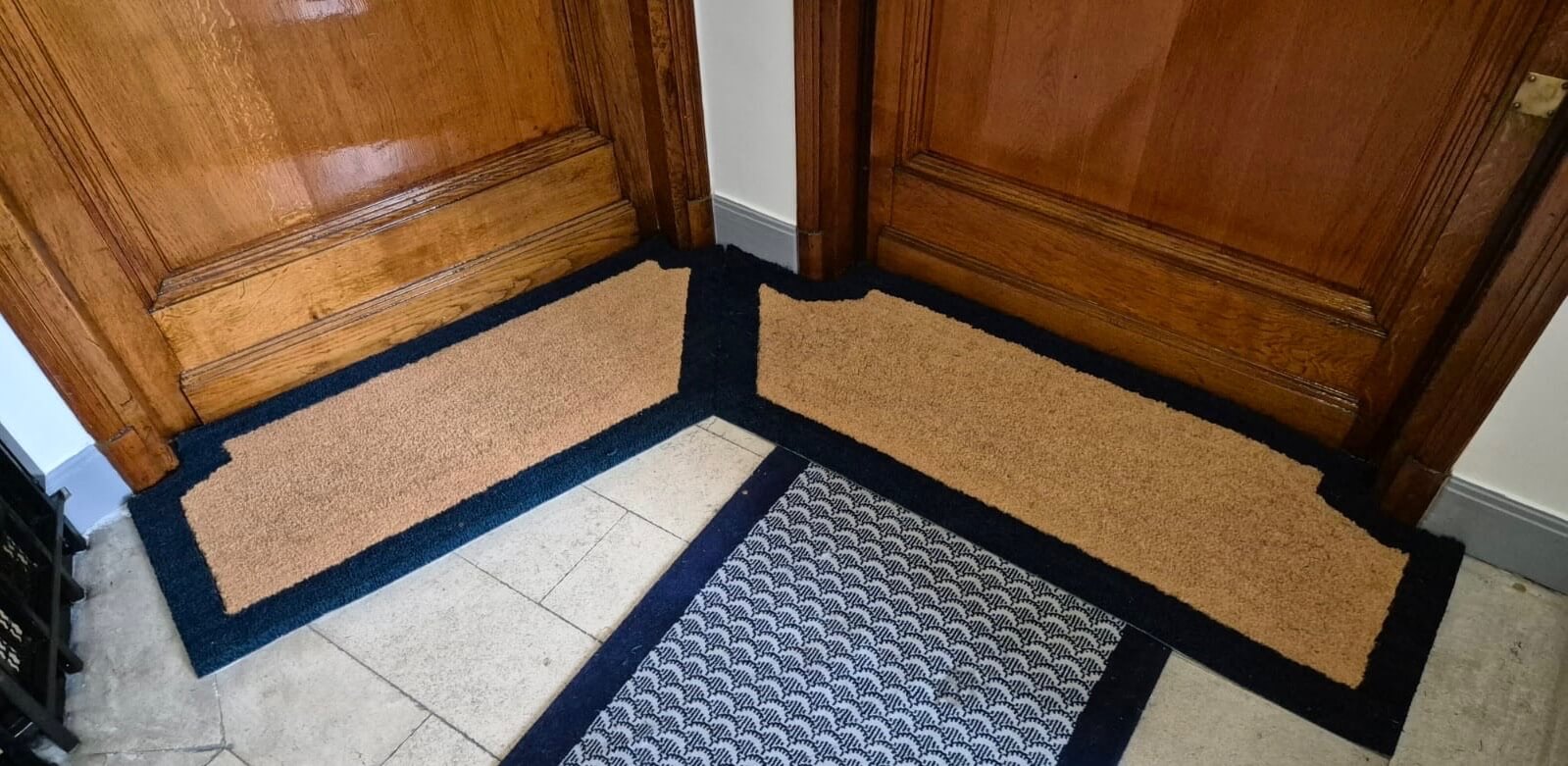 Deux paillassons d'entrée en coin avec cadre noir devant des portes en bois foncé.