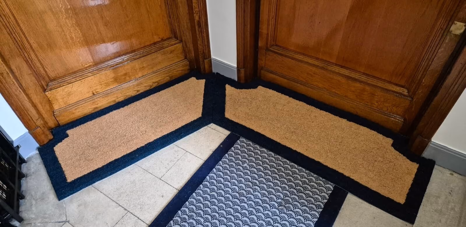Deux paillassons d'entrée en coin avec cadre noir devant des portes en bois foncé.
