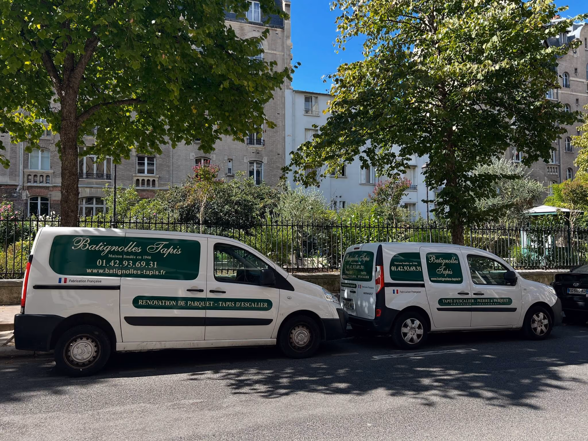 Deux camionnettes blanches Batignolles Tapis stationnées sous arbres le long d'une rue avec bâtiments en arrière-plan.