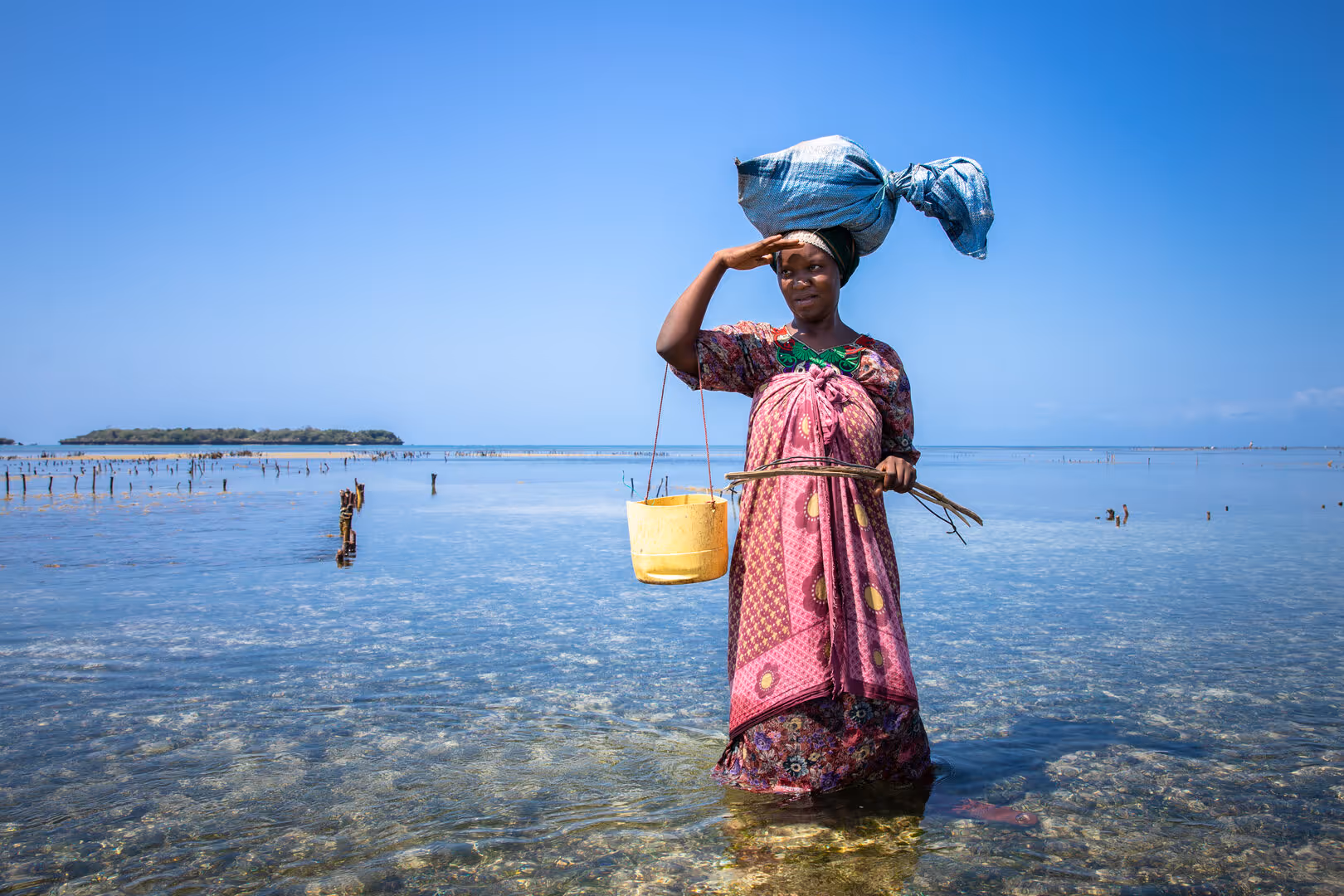 Woman standing in shallow clear water, carrying a bundle on her head and holding a yellow bucket.