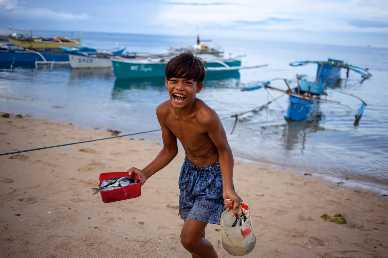 Boy laughing on beach holding a container and bucket filled with fish with boats in background.