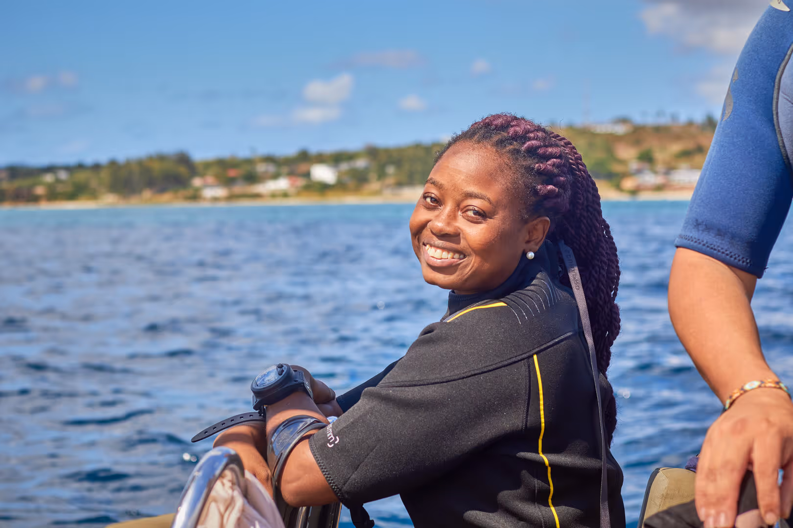Smiling woman with braided hair in a wetsuit on a boat with water and shoreline in the background.