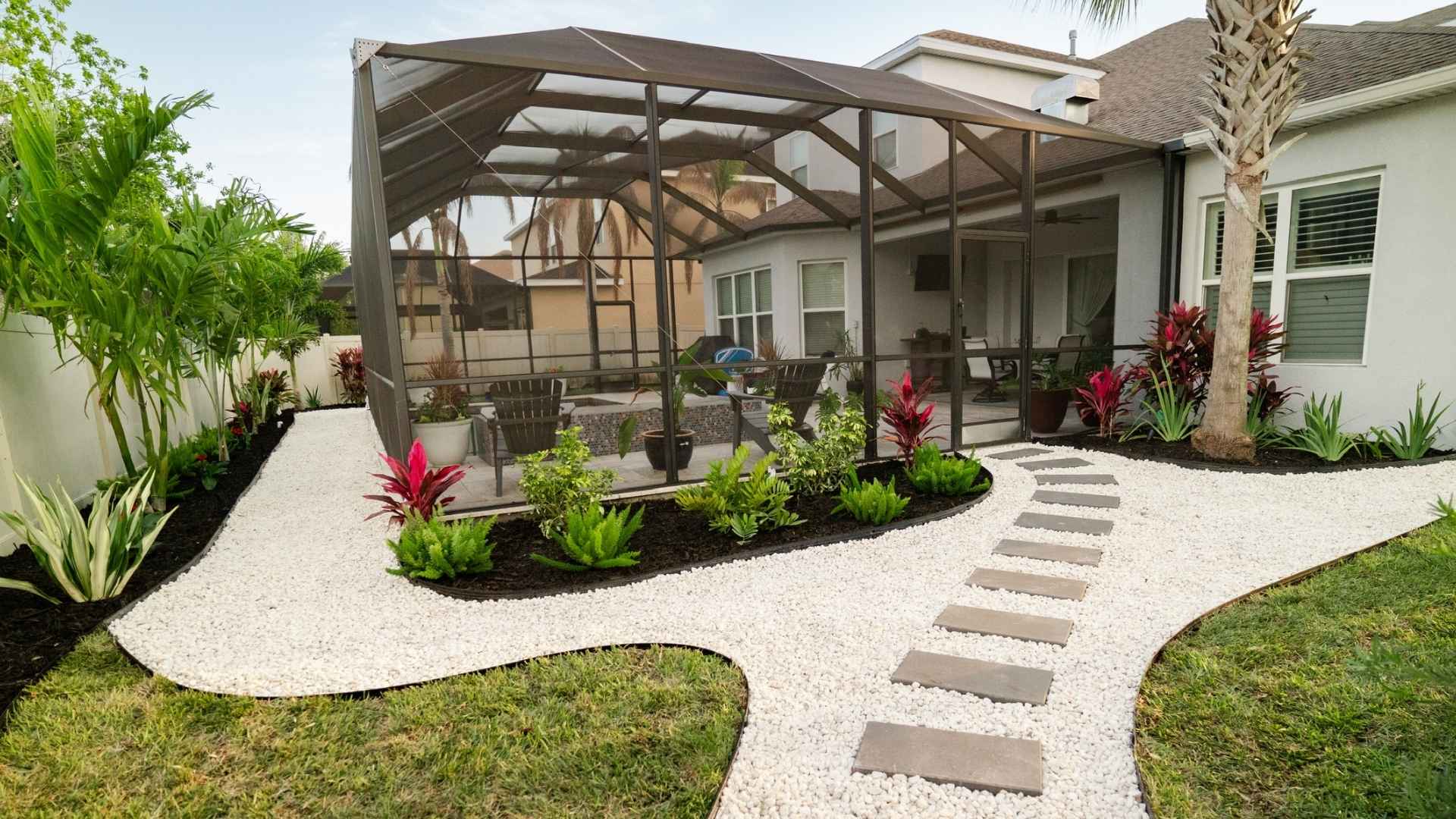 Completed backyard landscape with tropical plantings, white decorative stone, and a stepping-stone path surrounding a screened patio