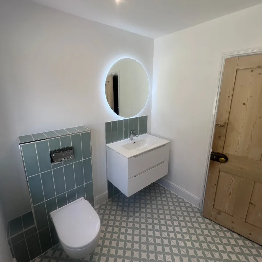Modern bathroom vanity unit with illuminated round mirror, wall-mounted basin, concealed toilet cistern and green feature tiles.