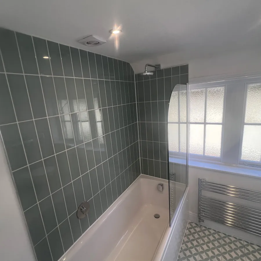 Modern bathroom renovation featuring sage green subway tiles, white bath with glass shower screen and patterned floor tiles.