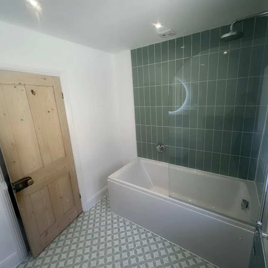 Finished bathroom refurbishment with white bath, sage green tiled shower wall, timber door and decorative patterned floor.
