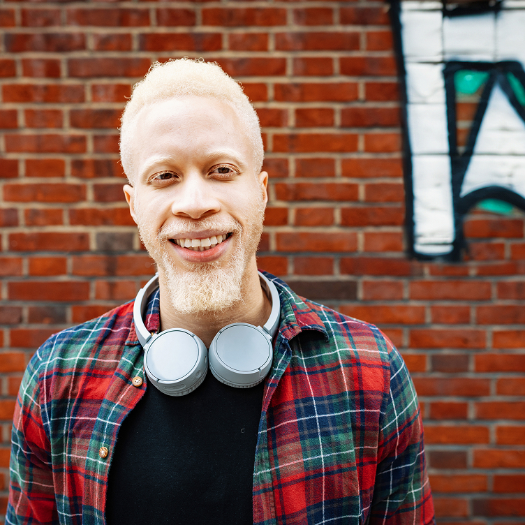 Smiling young man with blond hair and beard wearing a plaid shirt and gray headphones around neck.
