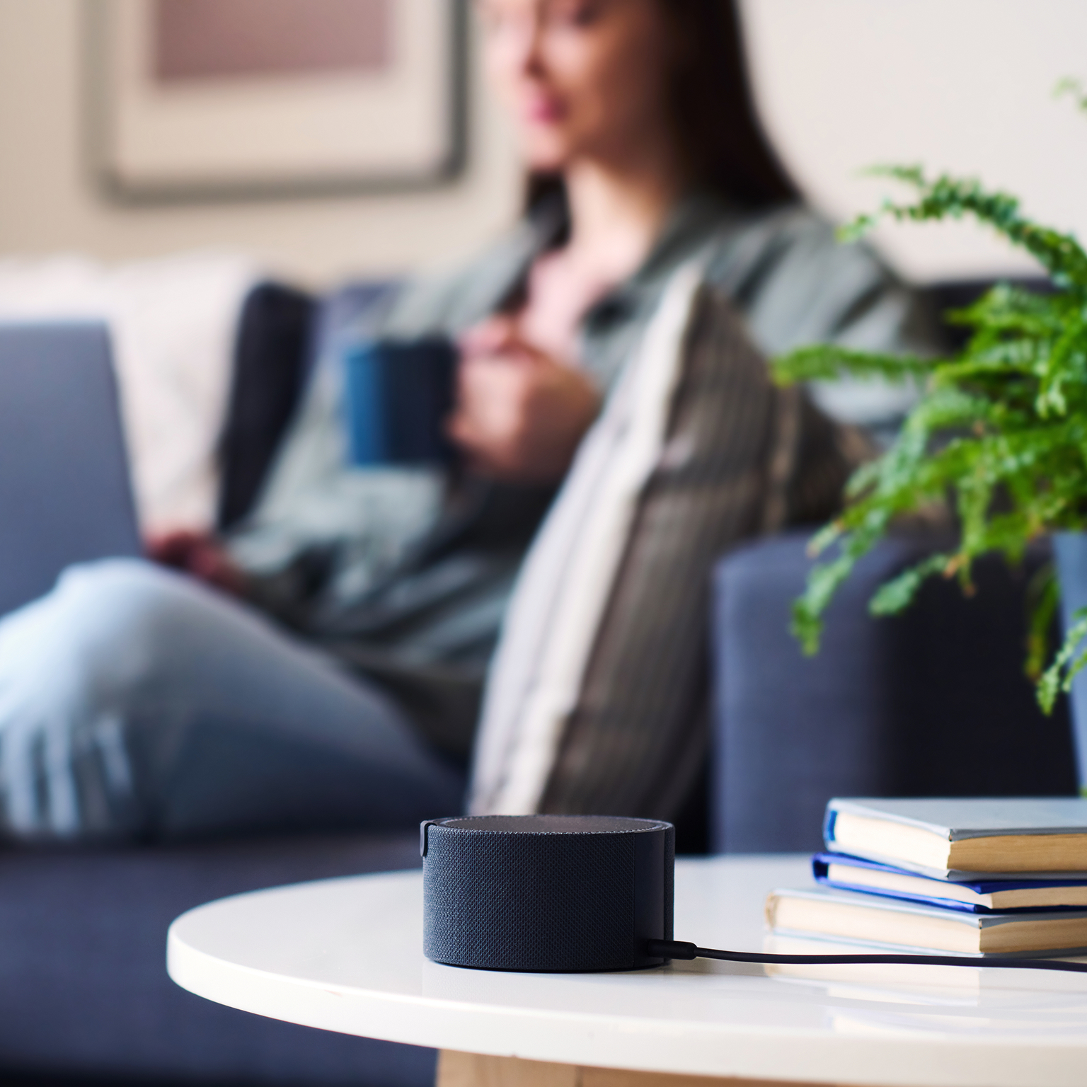 Black smart speaker on table with books and a blurred woman holding a cup on couch behind.