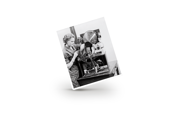 Black and white photo of a woman adjusting wires on a vintage scientific apparatus on a table.