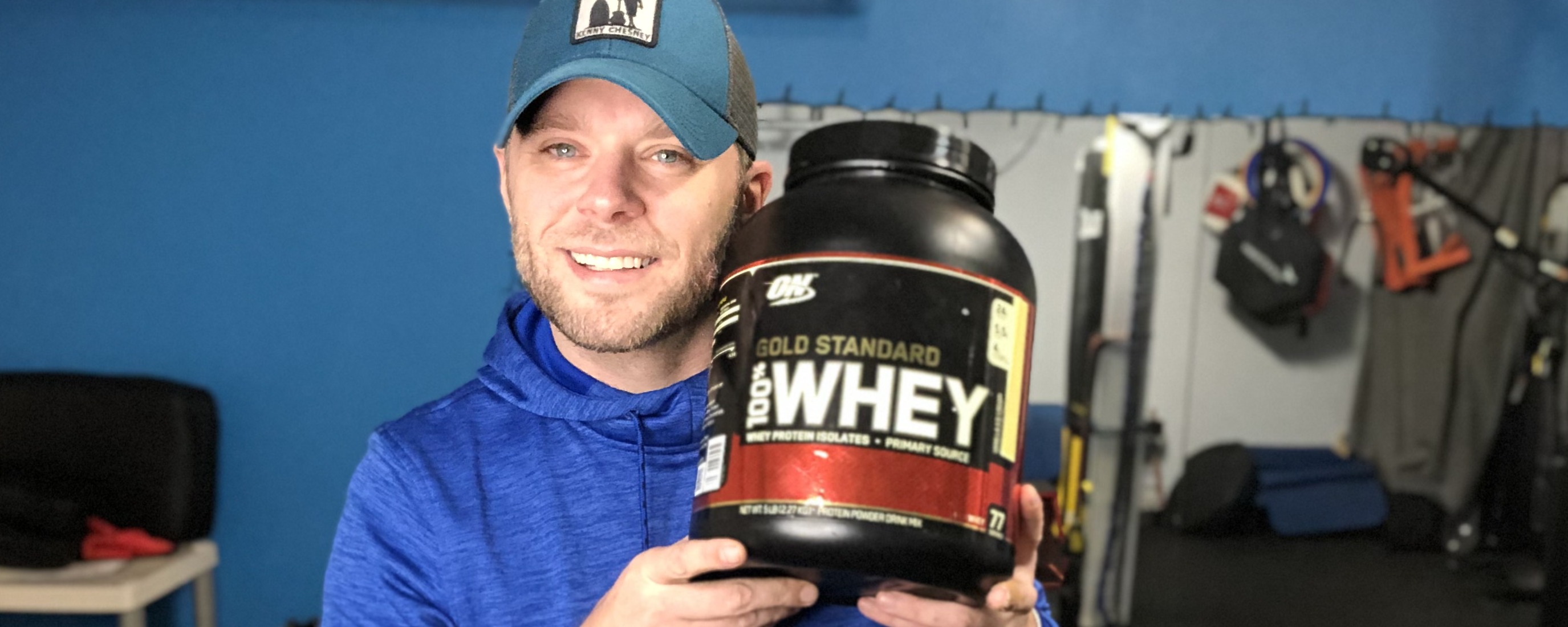 Corey holding a 5 lb tub of Optimum Nutrition Gold Standard 100 percent Whey Protein in the gym