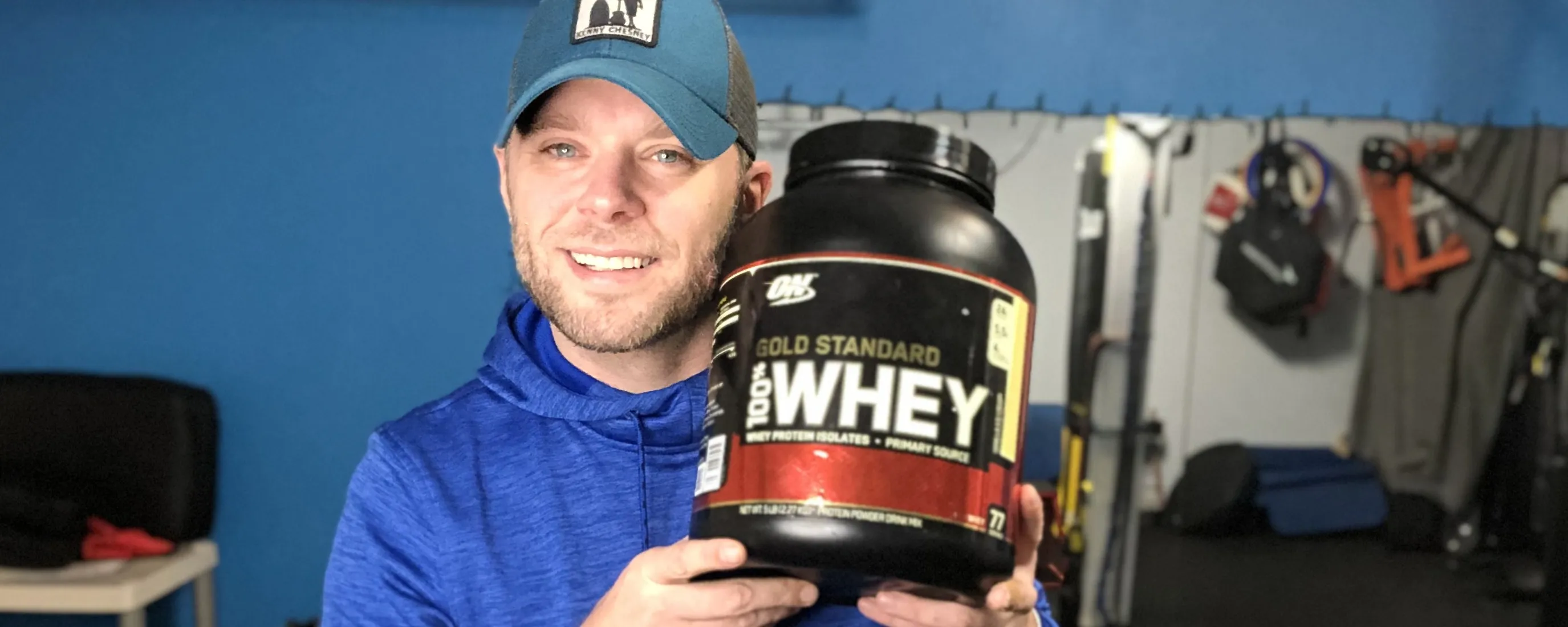 Corey holding a 5 lb tub of Optimum Nutrition Gold Standard 100 percent Whey Protein in the gym