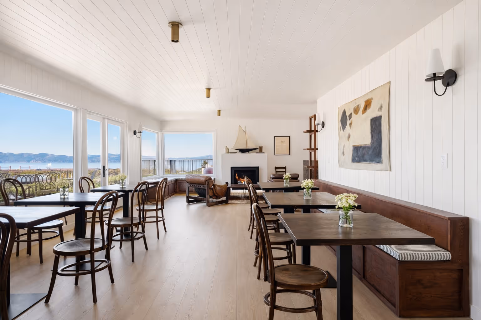 Bright café with wooden tables and chairs, fireplace, large windows, and a view of mountains and water.