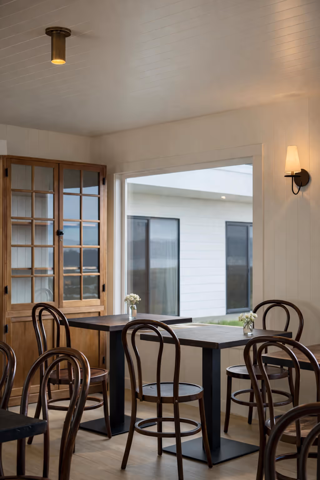 Minimalist dining area with wooden chairs, black tables, a wooden cabinet, and small flower vases on tables.