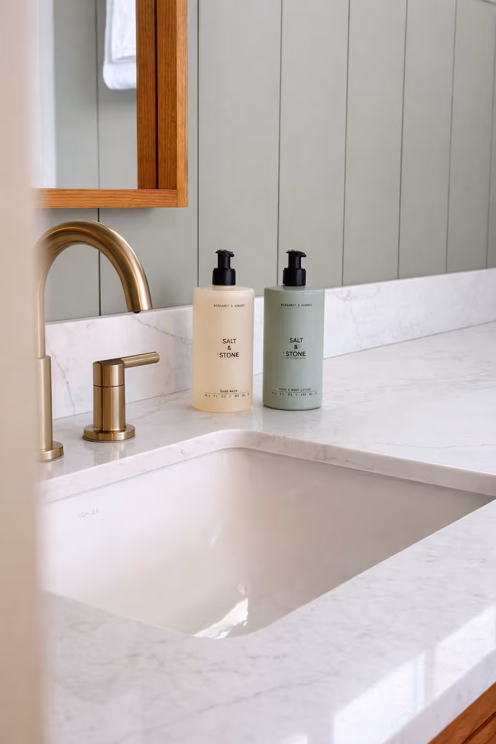 Modern bathroom sink with brass faucet and two Salt & Stone soap dispenser bottles on white marble countertop.
