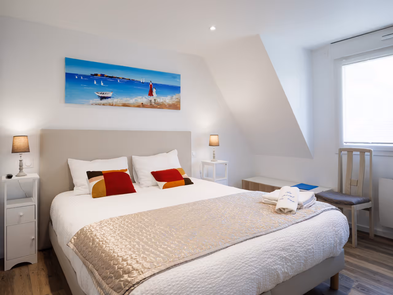 Chambre lumineuse avec grand lit double, tables de chevet, lampe, textile beige et tableau marin au mur.