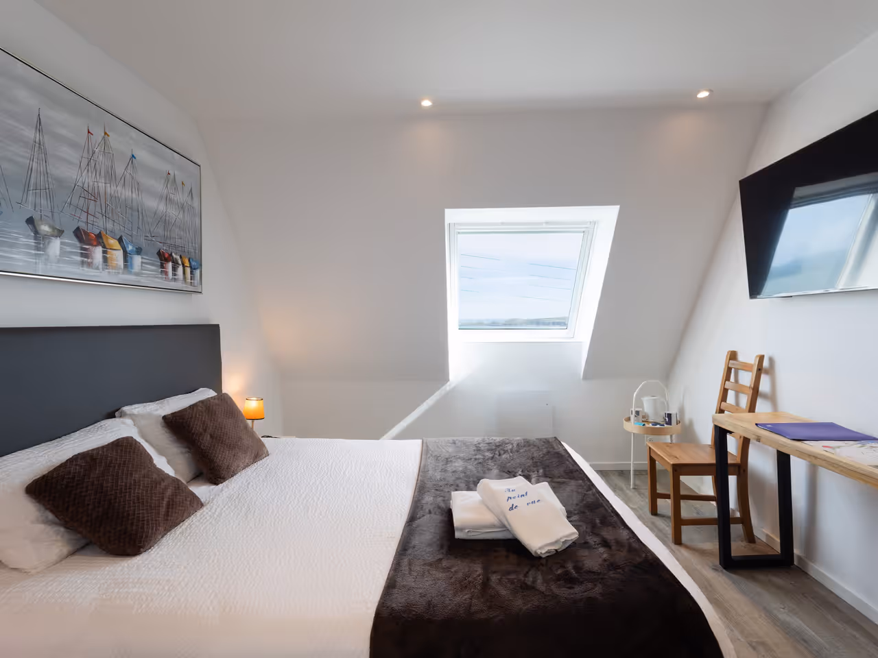 Chambre moderne avec lit double, coussins marron, bureau en bois, chaise et fenêtre au plafond.