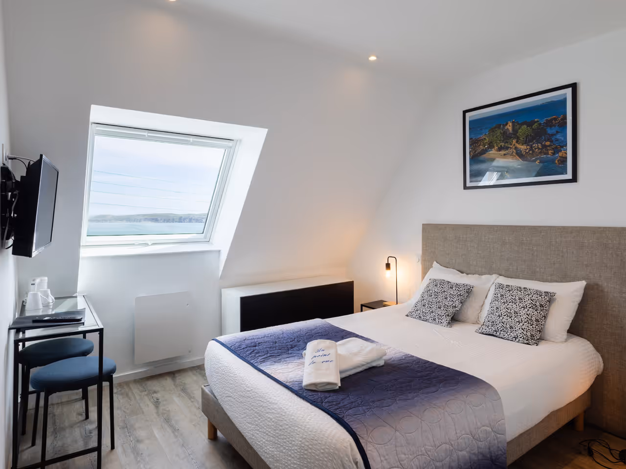 Chambre lumineuse avec lit double, table, TV murale et fenêtre inclinée donnant sur la mer.