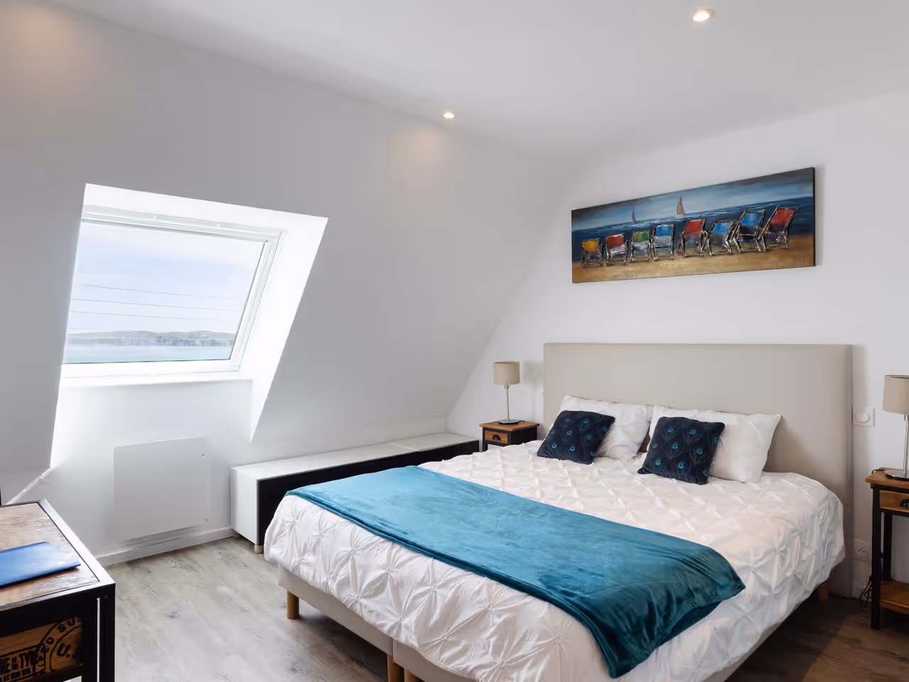 Chambre moderne avec grand lit double, coussins bleus, fenêtre inclinée et vue sur la mer.
