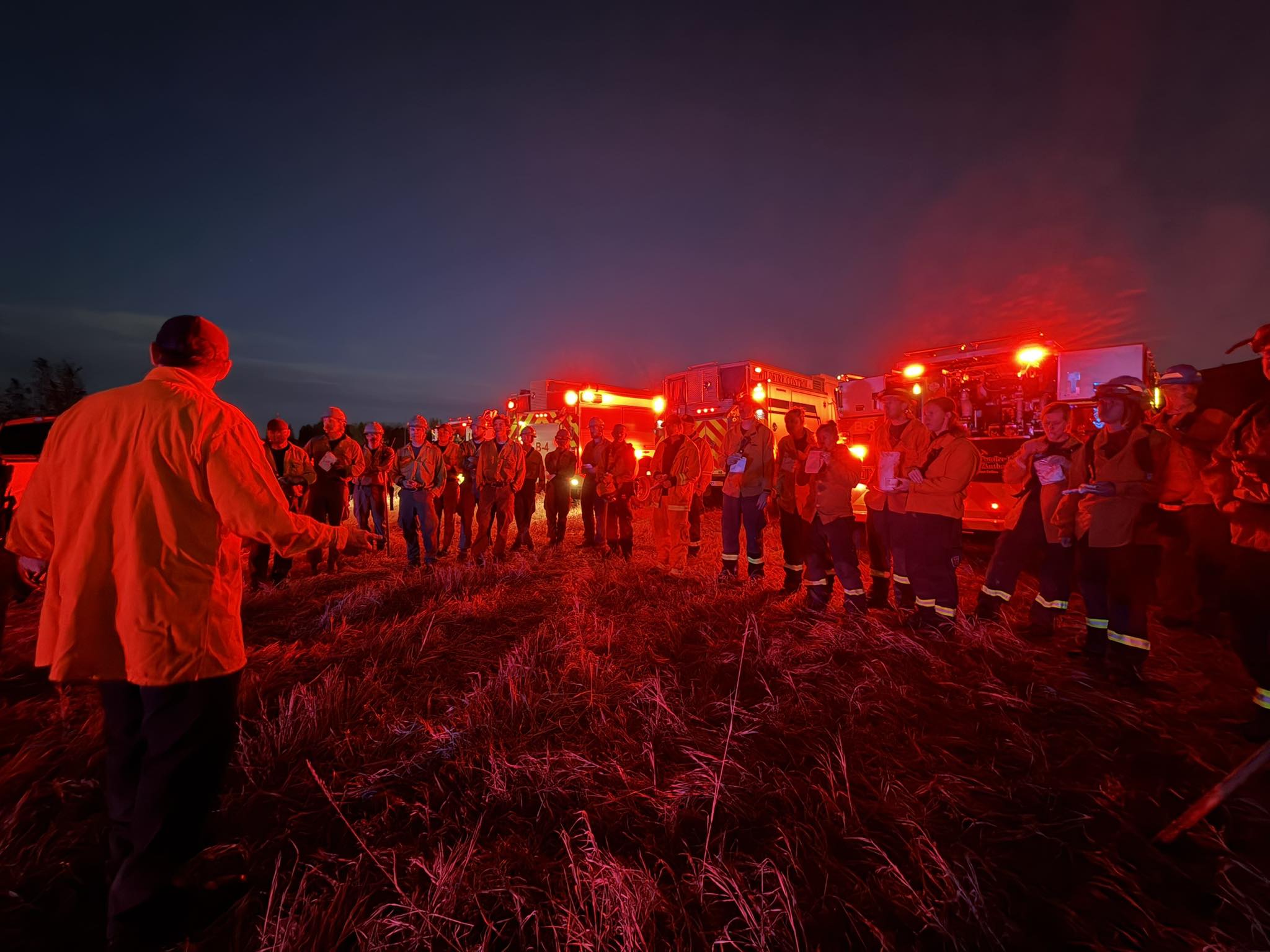 Firefighters in orange gear gather at night near fire trucks with red emergency lights on.