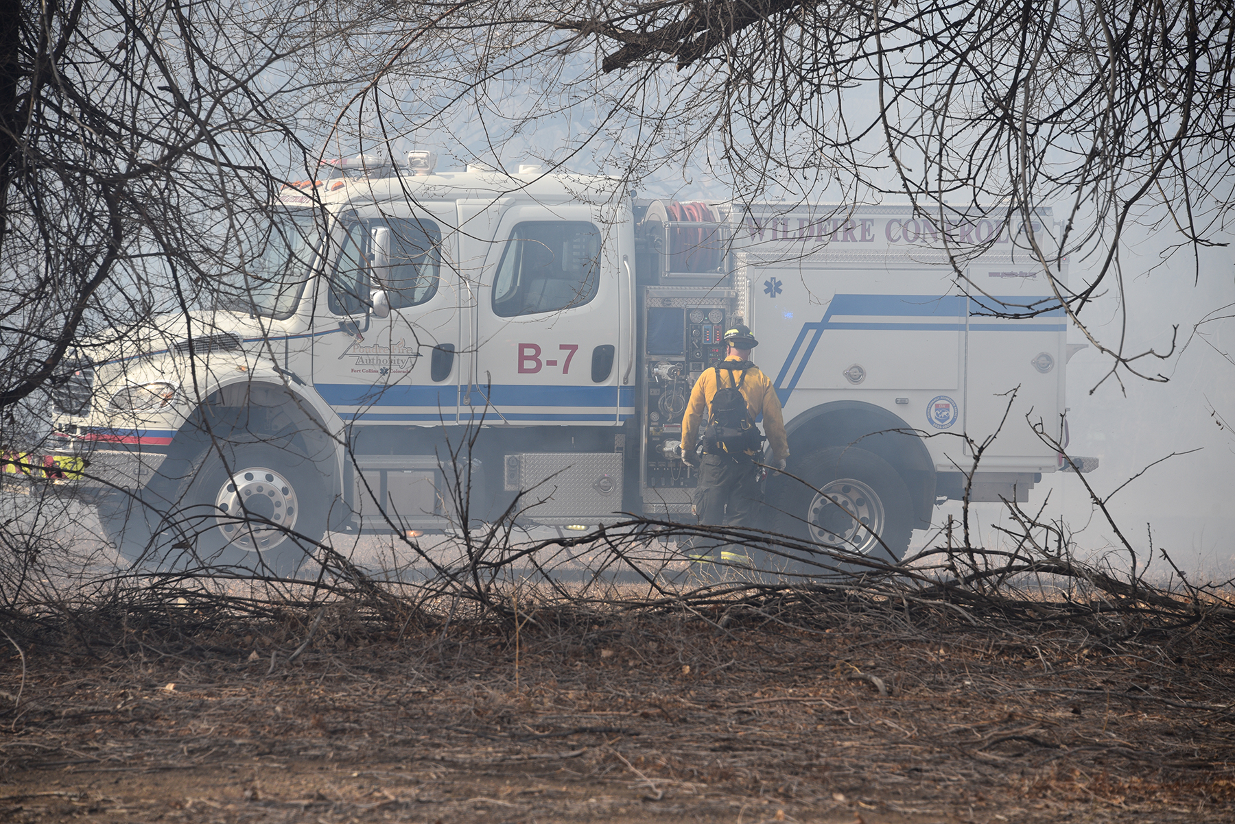 Firefighter in yellow jacket standing next to a white wildfire control truck in smoky area.