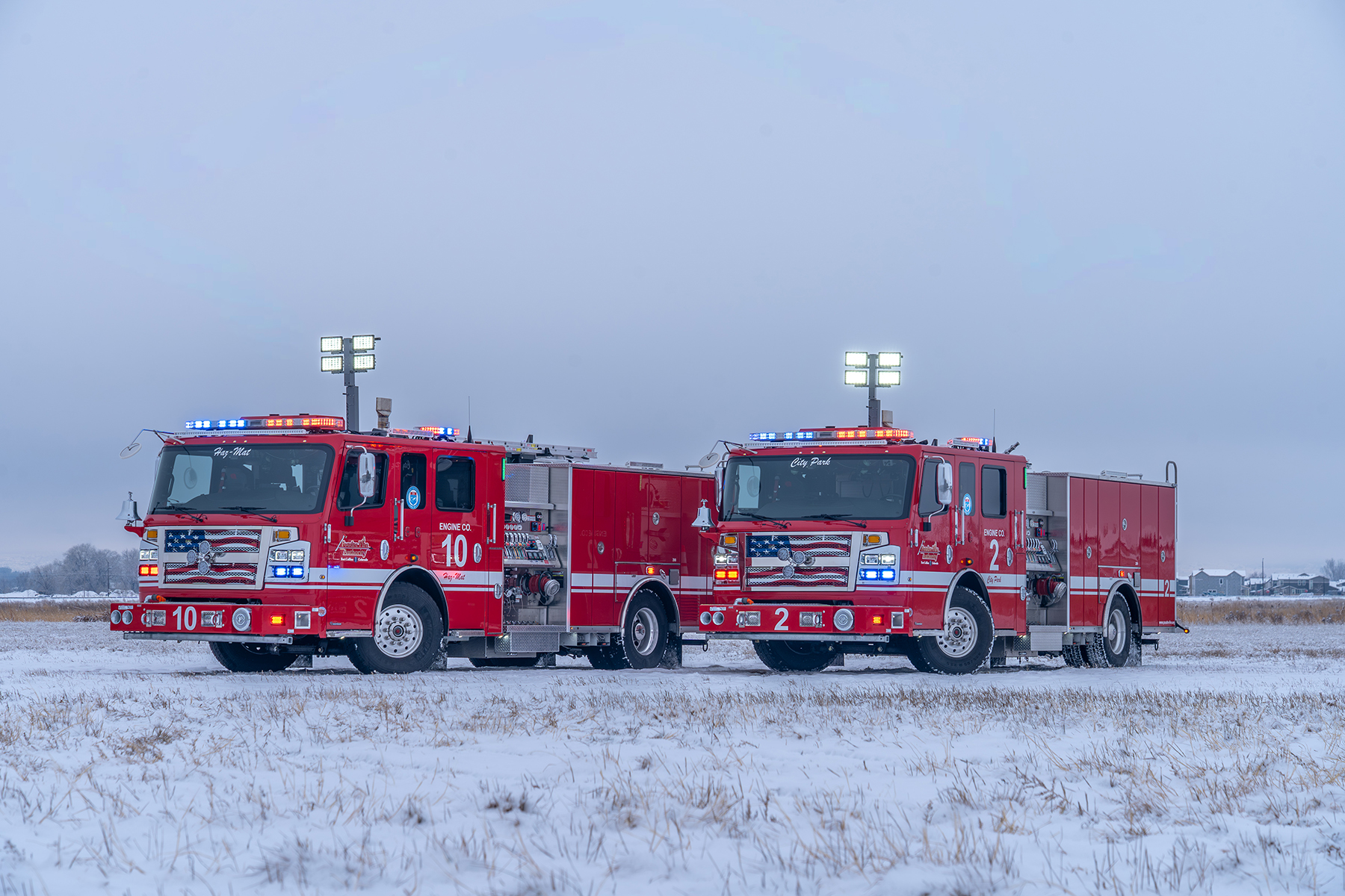 Two red fire trucks with flashing lights parked on snowy field under a cloudy sky.