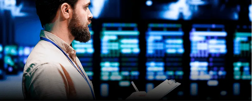 Man with beard holding clipboard and pen, looking at digital data screens in a control room.