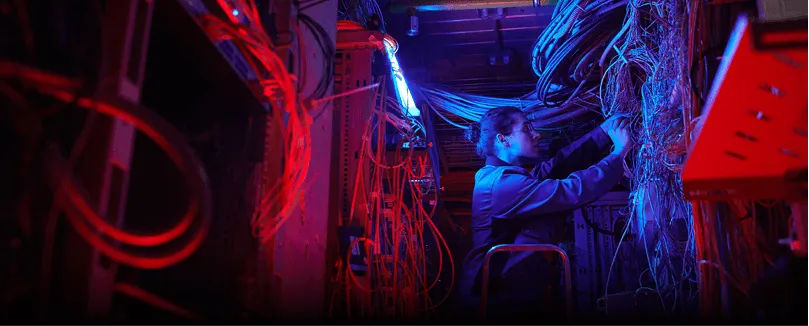 Technician working on complex network cables inside a server room lit with red and blue lights.