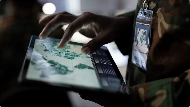 Person in camouflage uniform using a touchscreen tablet displaying a map with data panels.