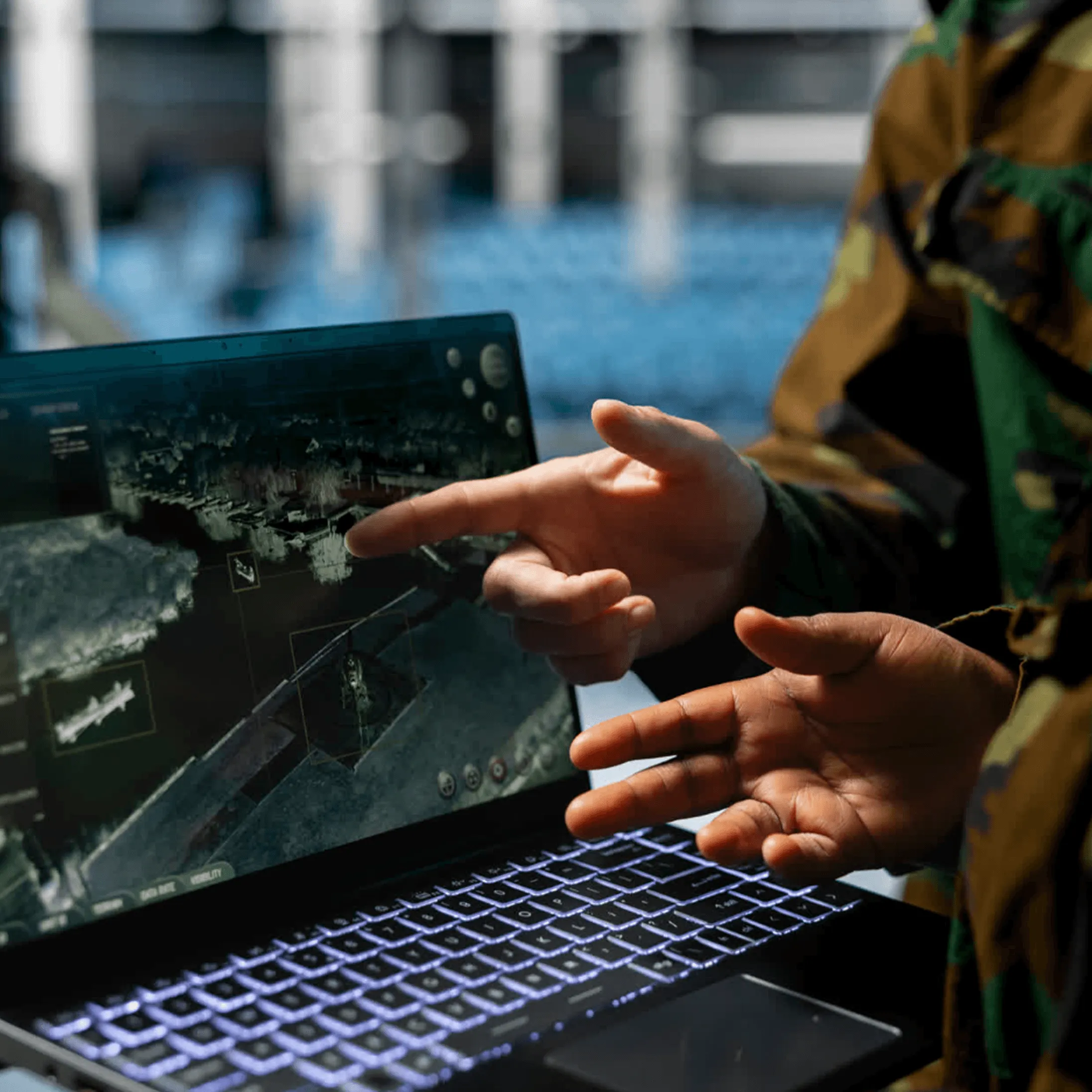 Two hands in camouflage pointing at a tactical map displayed on a laptop screen with backlit keyboard.