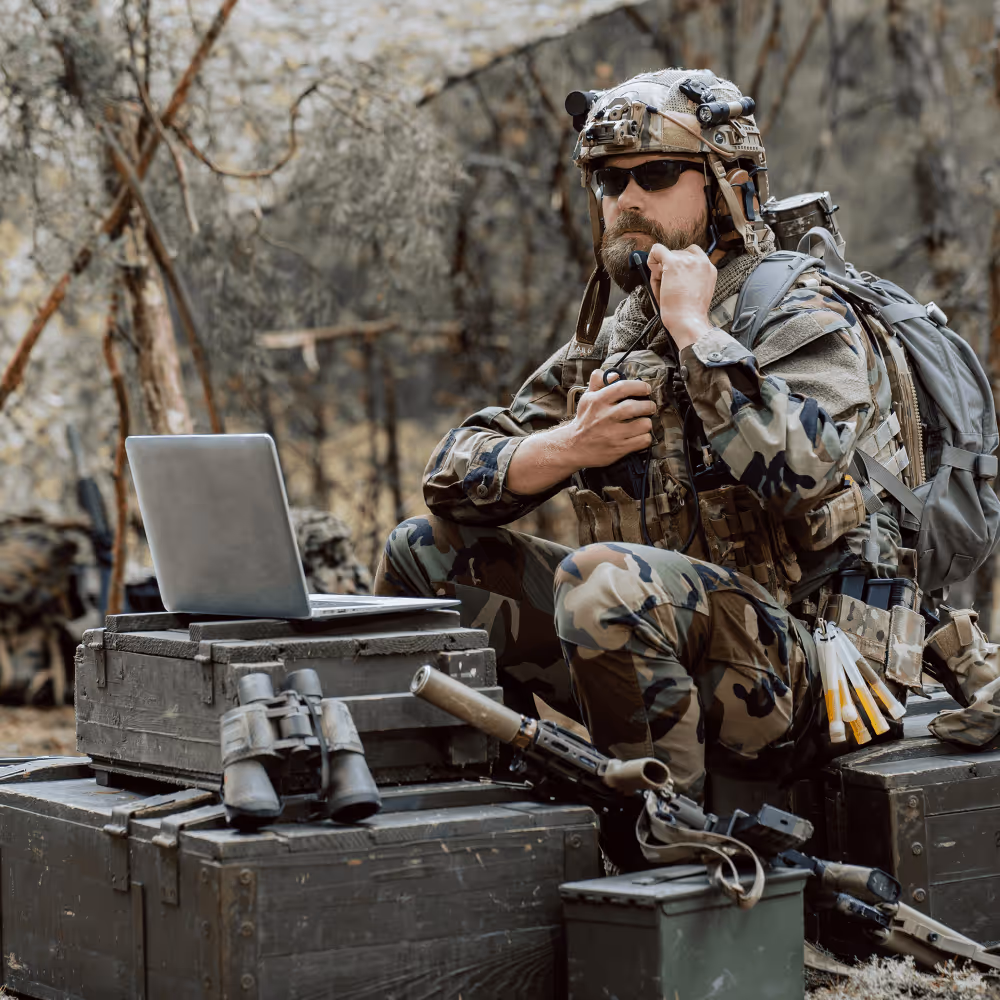Soldier in camouflage gear with helmet and sunglasses talks on radio near laptop and binoculars outdoors.