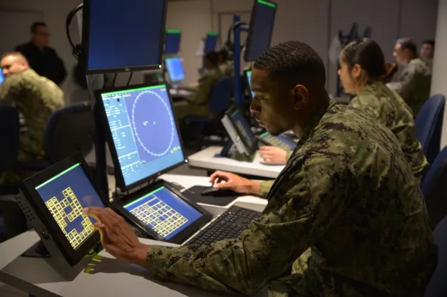 Military personnel operating radar and touchscreen displays in a control room environment.