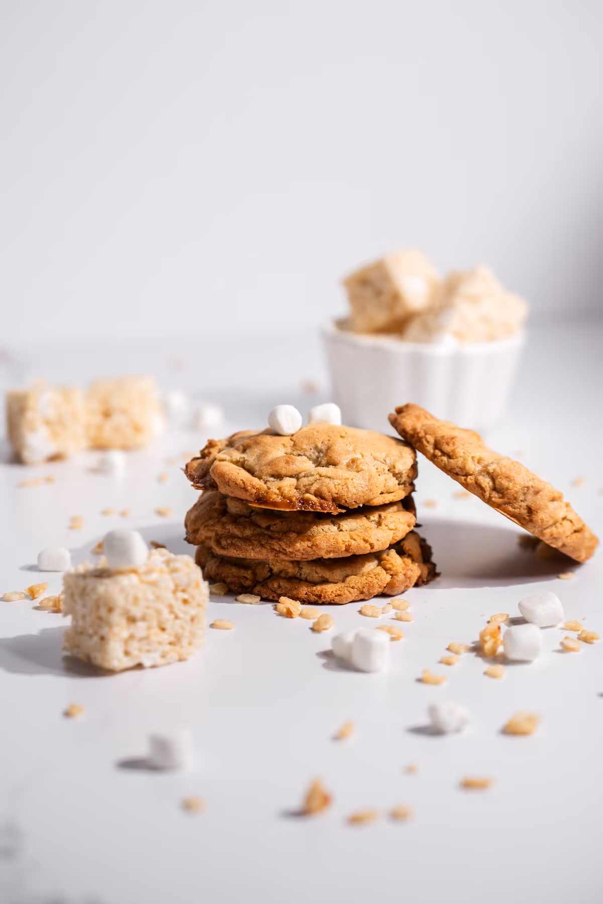 Stack of chewy cookies with mini marshmallows and rice Krispies treats scattered on white surface.
