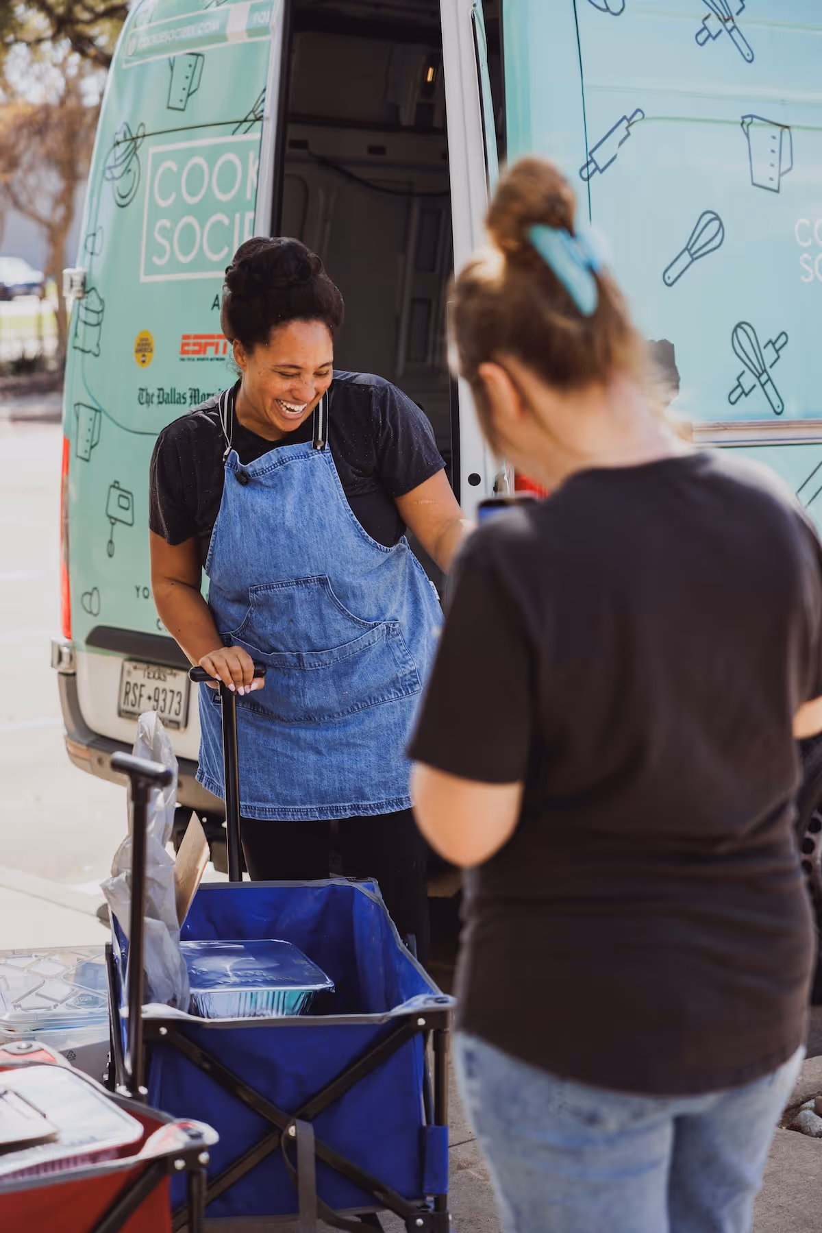 Woman in denim apron smiling by a van with food containers in a blue cart, another woman in foreground.