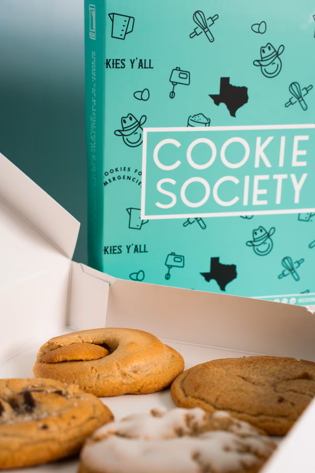 Close-up of assorted cookies in an open box with a turquoise Cookie Society package in background.