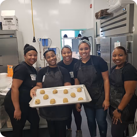 Five women smiling in kitchen, one holding tray with cookie dough balls ready to bake.