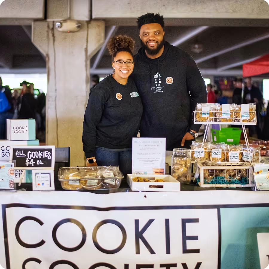Two smiling people behind a Cookie Society table selling packaged cookies at a market or event.