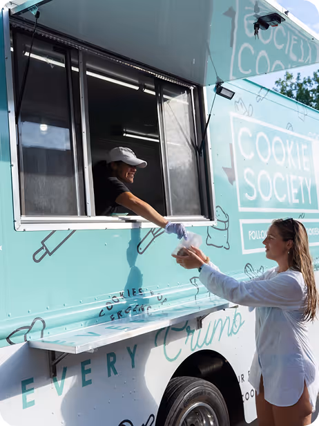 Woman receives food from smiling man inside a teal Cookie Society food truck window.
