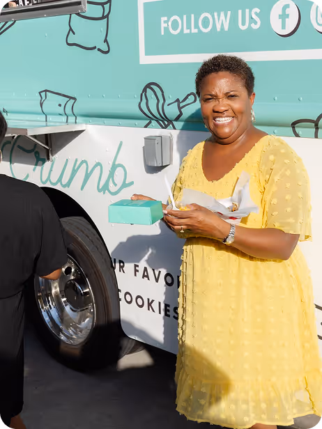 Smiling woman in yellow dress holds a teal box and eats near a food truck.