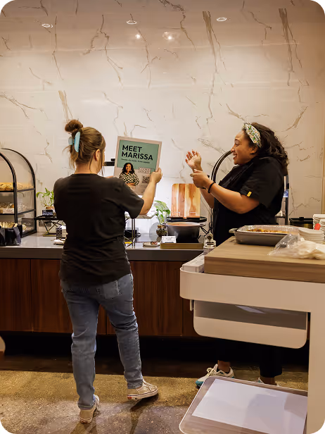 Two women in a kitchen, one holding a Meet Marissa poster, the other gesturing excitedly.