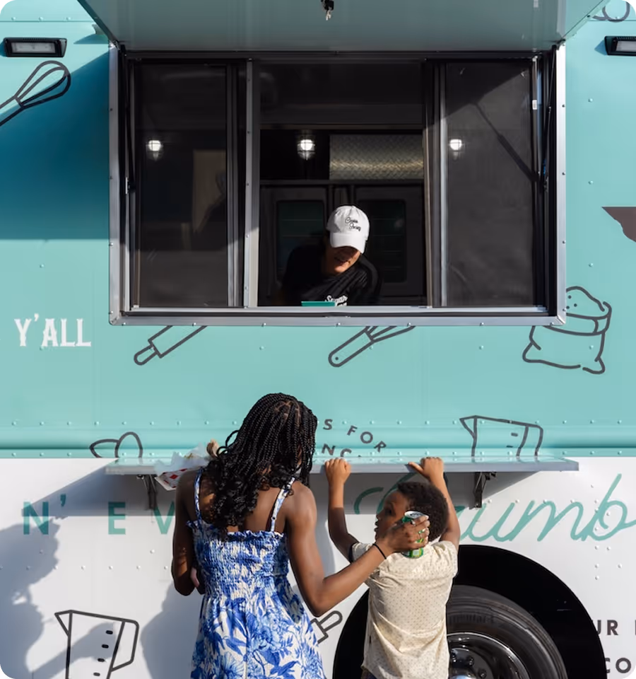 Two children stand at a teal food truck window where a person wearing a white cap serves them.
